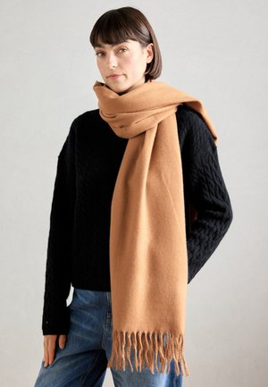 Écharpe en laine brune avec des franges, drapée autour d'un pull noir à texture, mettant en valeur sa douceur et sa couleur chaude. Un bas en denim décontracté complète le look.