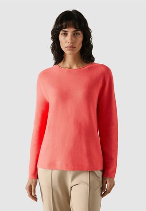 Mujer con cabello largo y ondulado oscuro, vistiendo un suéter coral de manga larga y pantalones beige, mirando hacia adelante contra un fondo liso de color gris claro.