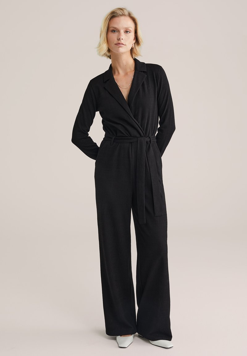 Melnā kombinezonā ar garām piedurknēm, apkakli un jostu viduklī. Izgatavots no teksturēta auduma, ar platām biksēm.