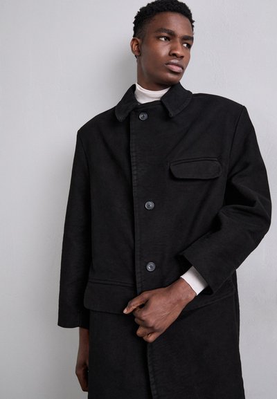 Black, button-up coat with a collar, chest pocket, and long sleeves. Made of textured material, featuring a clean design and simple lines.