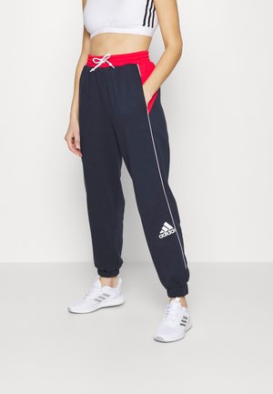 Kobieta w granatowych dresach z czerwoną gumą w pasie, białym sznurkiem i logo Adidas, zestawionych z białymi trampkami i białym krótki topem.
