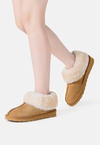 Tan suede slippers with a fluffy cream-colored faux fur cuff, rounded toe, and textured rubber sole. No visible branding on the exterior.