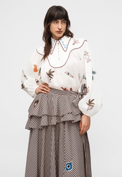 Γυναίκα που φοράει λευκό πουκάμισο με πολύχρωμη αφηρημένη κεντητή διακόσμηση και καφέ ριγέ φούστα με στρώσεις δεμένη στη μέση, στέκεται μπροστά σε απλό φόντο.