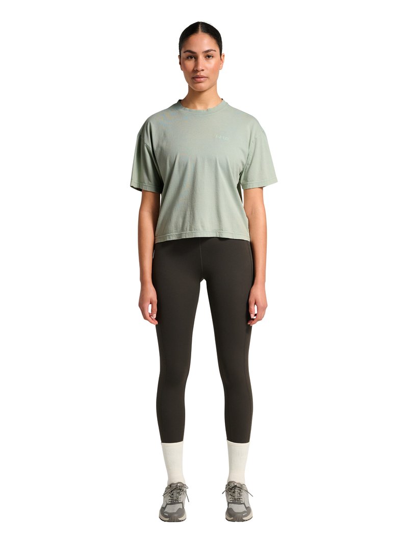 Kobieta stoi zwrócona przodem, ubrana w jasnokrótki zielony oversize t-shirt, czarne legginsy, białe skarpetki i szare trampki na białym tle.