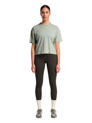 Kobieta stoi zwrócona przodem, ubrana w jasnokrótki zielony oversize t-shirt, czarne legginsy, białe skarpetki i szare trampki na białym tle.