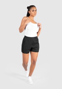 Schwarze Sportshorts mit elastischem Bund und Seitentaschen, kombiniert mit einem weißen Tanktop und weißen Sneakers, vor einem grauen Hintergrund.