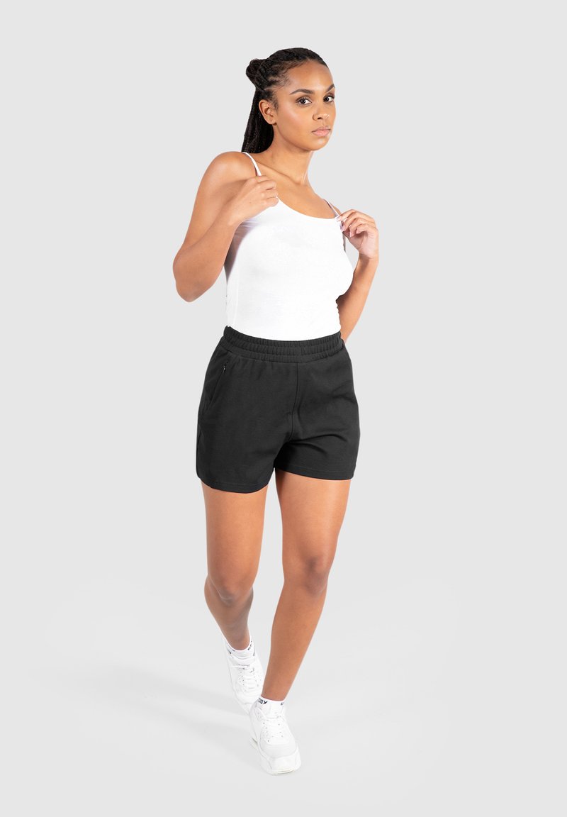 Zwarte sportshorts met een elastische tailleband en zijzakken, gecombineerd met een wit hemdtop en witte sneakers, tegen een grijze achtergrond.
