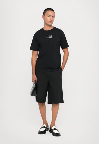 T-shirt en coton noir à manches courtes avec un design graphique sur la poitrine, associé à un short noir jusqu'aux genoux et des mocassins noirs brillants.
