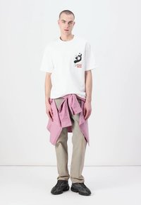 Maglietta bianca in cotone con stampa grafica sul lato sinistro del petto, abbinata a pantaloni beige e una camicia rosa a bottone annodata in vita. Sneakers nere.