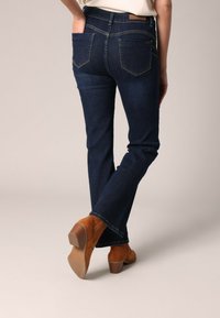 Jeans en denim à jambe évasée de couleur bleu foncé avec une texture lisse, dotés de deux poches arrière et d'une taille mi-haute, associés à des bottines ouvertes tan.