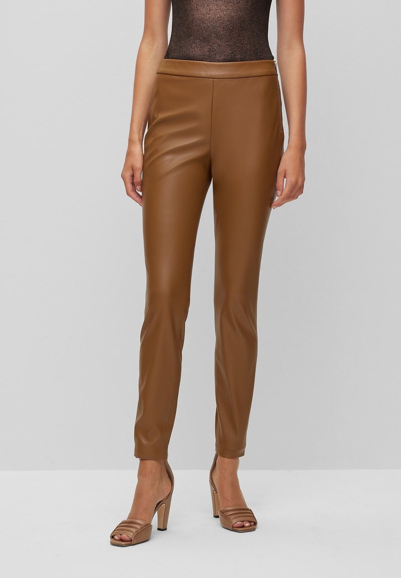 Bruna imitationläderbyxor med en åtsittande design, slät textur och elastisk midja, tillsammans med bronzfärgade högklackade sandaler.