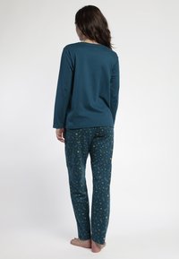 Mujer con cabello largo y castaño, de pie descalza y de espaldas, vistiendo una blusa de manga larga en color verde azulado y pantalones a juego con un estampado celestial dorado.