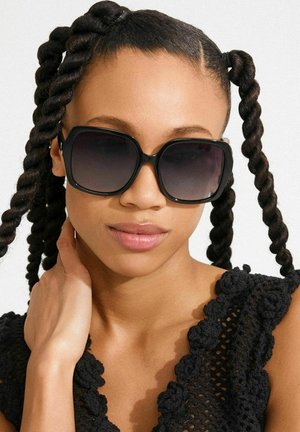 Black oversized sunglasses with gradient lenses, rectangular shape, and thick glossy frames. Model features braided hairstyles and wears a black textured top.