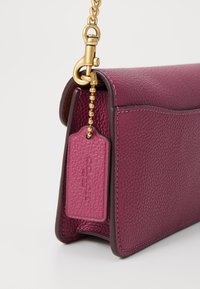 Sac bandoulière en cuir violet avec texture grainée, doté d'une chaîne en or et d'une étiquette suspendue avec un logo en relief.