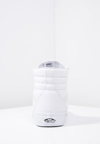 White high-top sneakers made of canvas with padded collar, featuring a black logo tag on the heel and subtle stitching details.