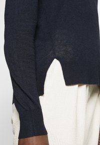 Granatowy sweter dzianinowy z prążkowanymi mankietami, z detalem w postaci rozcięcia na dole. Projekt jest lekki o miękkiej fakturze.