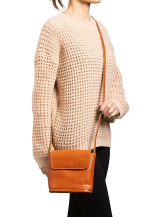 Femme portant un pull beige en maille et un pantalon noir, avec un sac bandoulière en cuir marron clair à rabat.