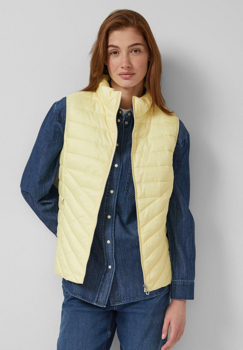 Frau trägt eine hellgelbe gesteppte Weste über einem dunkelblauen Jeanshemd mit Knöpfen sowie passende Jeanshosen und steht vor einem einfarbigen Hintergrund.