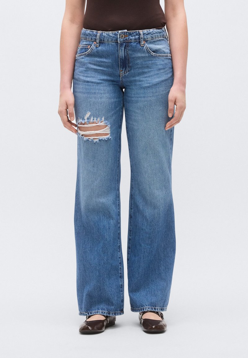Blauwe jeans met wijde pijpen gemaakt van denim. Heeft een groot distressed gat op de linkerdij. Klassiek ontwerp met vijf zakken en riemlussen.