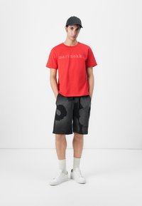 Maglietta rossa in cotone con la scritta "marimekko", pantaloni corti neri a fantasia, sneakers bianche e calzini grigi chiari, su uno sfondo semplice.