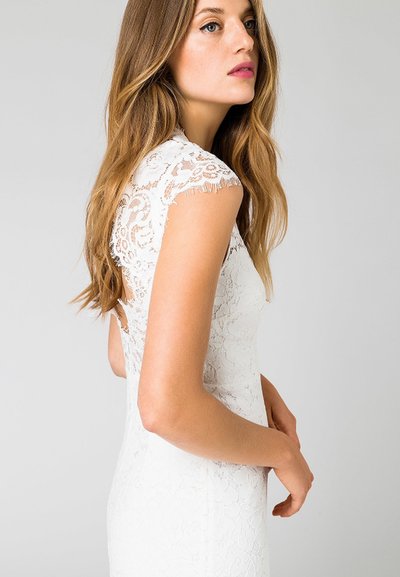 Λευκό φόρεμα με δαντέλα και κοντά μανίκια, που διαθέτει λουλουδάτα σχέδια και εφαρμοστή γραμμή, τονίζοντας την περίπλοκη υφή και την ομαλή επιφάνεια.