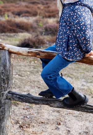 Stivaletti neri con suole texturizzate, jeans in denim blu e una giacca blu a fantasia floreale con cappuccio. Seduto su una recinzione di legno.