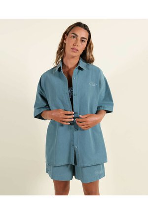 Femme portant une chemise boutonnée surdimensionnée à manches courtes couleur sarcelle et un short assorti, ajustant les boutons de la chemise, fond neutre.