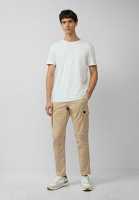 Biała koszulka z krótkim rękawem, beżowe spodnie cargo z bocznymi kieszeniami i białe sportowe buty z szarymi akcentami. Prosty, casualowy strój.