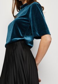 Tyrkysový sametový krátký rukáv top s vysokým límcem, kombinovaný s černou skladanou sukní. Tkanina vykazuje měkkou strukturu a jemný lesk.