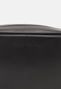 Černá texturovaná kožená taška s kulatým vrchem, vybavená stříbrným zipem a jemně raženým logem "Calvin Klein Jeans" na přední straně.