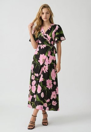 Mujer con cabello rubio largo que lleva un vestido cruzado negro con estampado floral rosa y sandalias negras de tacón alto con tiras, de pie sobre un fondo blanco.