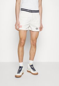 Bílé sportovní šortky s tmavě modrým pruhovaným pasem, dvěma růžovými pruhy po stranách a logem na levém stehně.