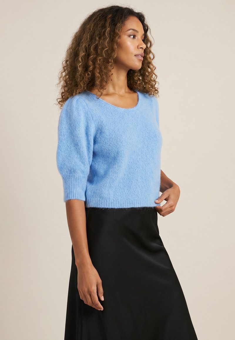 Femme aux cheveux bouclés portant un pull duveteux bleu clair et une jupe noire, debout sur un fond neutre, regardant sur le côté.