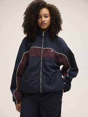Joven con cabello rizado, vestida con una chaqueta deportiva con cremallera en azul marino y burdeos y pantalones a juego, de pie con una mano en el bolsillo.
