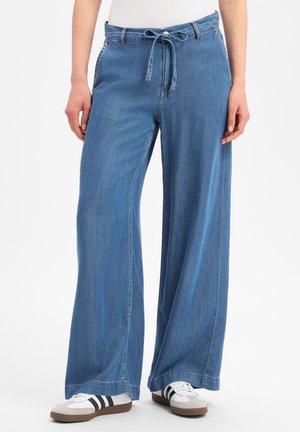 Pantalon en jean bleu à jambes larges avec une ceinture nouée à la taille, porté avec des baskets blanches à rayures noires sur une personne debout.