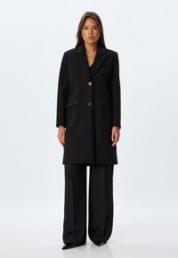 Abrigo de lana negro con corte a medida, solapa tipo muesca, dos bolsillos frontales y cierre de dos botones. Combinado con pantalones de pierna ancha negros.