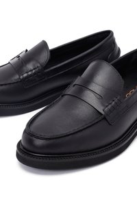 Mocasines de cuero negro con un acabado suave, detalles de costura y punta redondeada. Presentan una correa decorativa en la parte frontal.