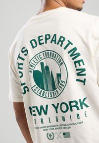 T-shirt en coton crème avec imprimé graphique vert incluant le texte "DÉPARTEMENT DE SPORTS" et "NEW YORK". Comprend un design de la ligne d'horizon de la ville et la date d'établissement.