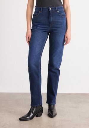 Mørkeblå high-waisted jeans med rett ben, som har fem lommer og subtile sømdetaljer, kombinert med svarte høyhælte ankelstøvler.