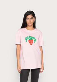 T-shirt de algodão rosa claro com texto verde "day dreaming" e um gráfico de morango vermelho com pontos brancos. Corte descontraído e mangas curtas.