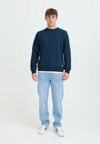 Sudadera azul marino de cuello redondo hecha de algodón, llevada sobre una camisa blanca. Combinada con jeans de color azul claro y zapatillas negras y blancas.