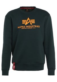 Sudadera verde de algodón con cuello redondo. Presenta un audaz texto y logotipo en naranja "ALPHA INDUSTRIES" en la parte delantera, con una etiqueta roja en el dobladillo.