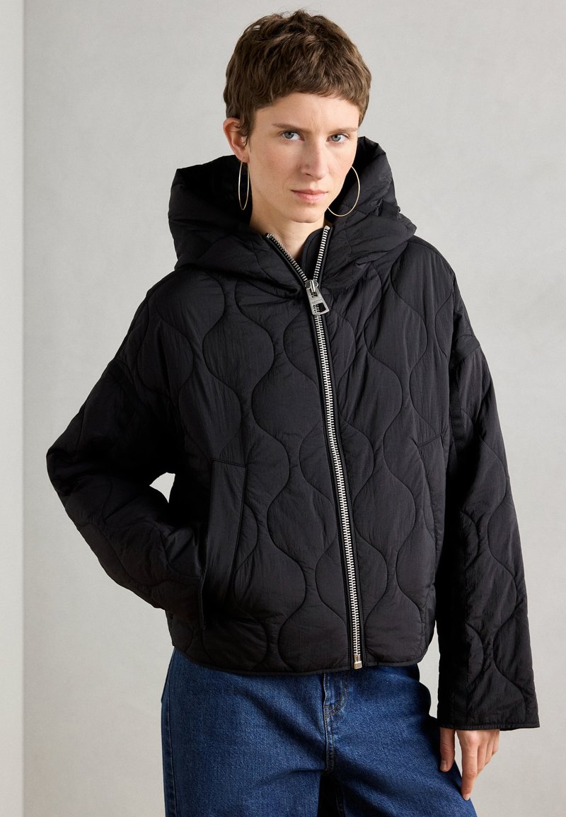 Veste matelassée noire avec capuche, présentant une fermeture éclair à l'avant, une silhouette oversize, et un motif texturé. Associée à un jean bleu foncé.