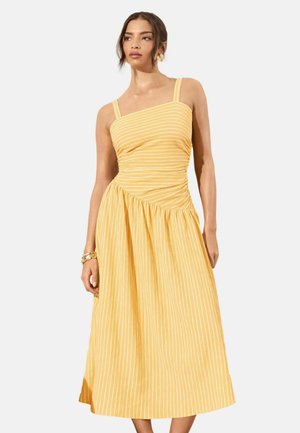 Femeie purtând o rochie midi galbenă fără mâneci, cu dungi verticale albe și o cusătură diagonală, accesorizată cu o brățară aurie.