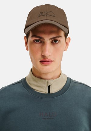 Junger Mann, der eine braune "Off Duty Gear"-Kappe und ein geschichtetes blau-beiges Reißverschluss-Oberteil trägt, schaut mit neutralem Gesichtsausdruck nach vorne.