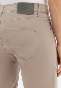 Beżowe spodnie z tylną kieszenią i naszywką z logo Brax, noszone przez osobę stojącą z prawą ręką widoczną przy boku.