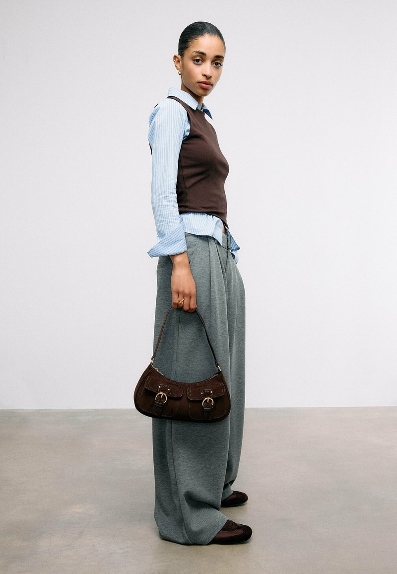 Femme portant une chemise rayée bleue avec un gilet marron, un pantalon large gris, tenant un sac à main marron, debout contre un mur blanc uni.