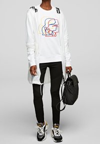 Camisa blanca de manga larga con detalles gráficos, combinada con pantalones negros ajustados. Accentuada con una bolsa negra y zapatillas deportivas negras y doradas.