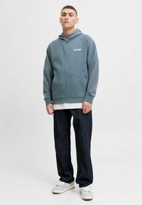 Haut à capuche zippé gris avec des accents côtelés, poches avant et logo. Porté avec un jean sombre ample et des baskets blanches. Texture lisse.