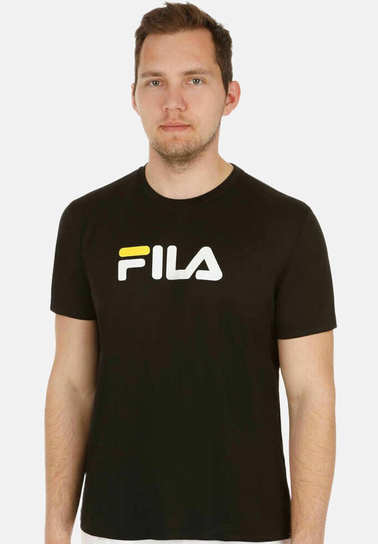 Man die een zwart FILA T-shirt draagt met een geel-wit logo, staand tegen een effen witte achtergrond.